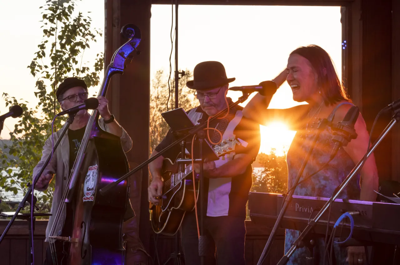 A three-piece band performs on an outdoor stage with a sunset in the background.