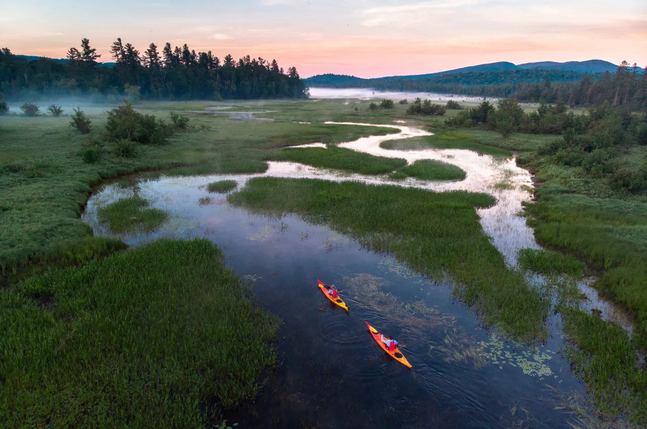 Two red kayaks paddle through an oxbow, a calm, bending river that loops back and forth amid shoreline plants.
