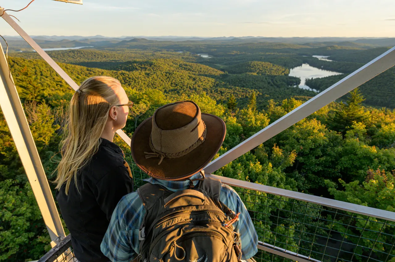 Two hikers look out over a vast, forested wilderness while the sun sets.