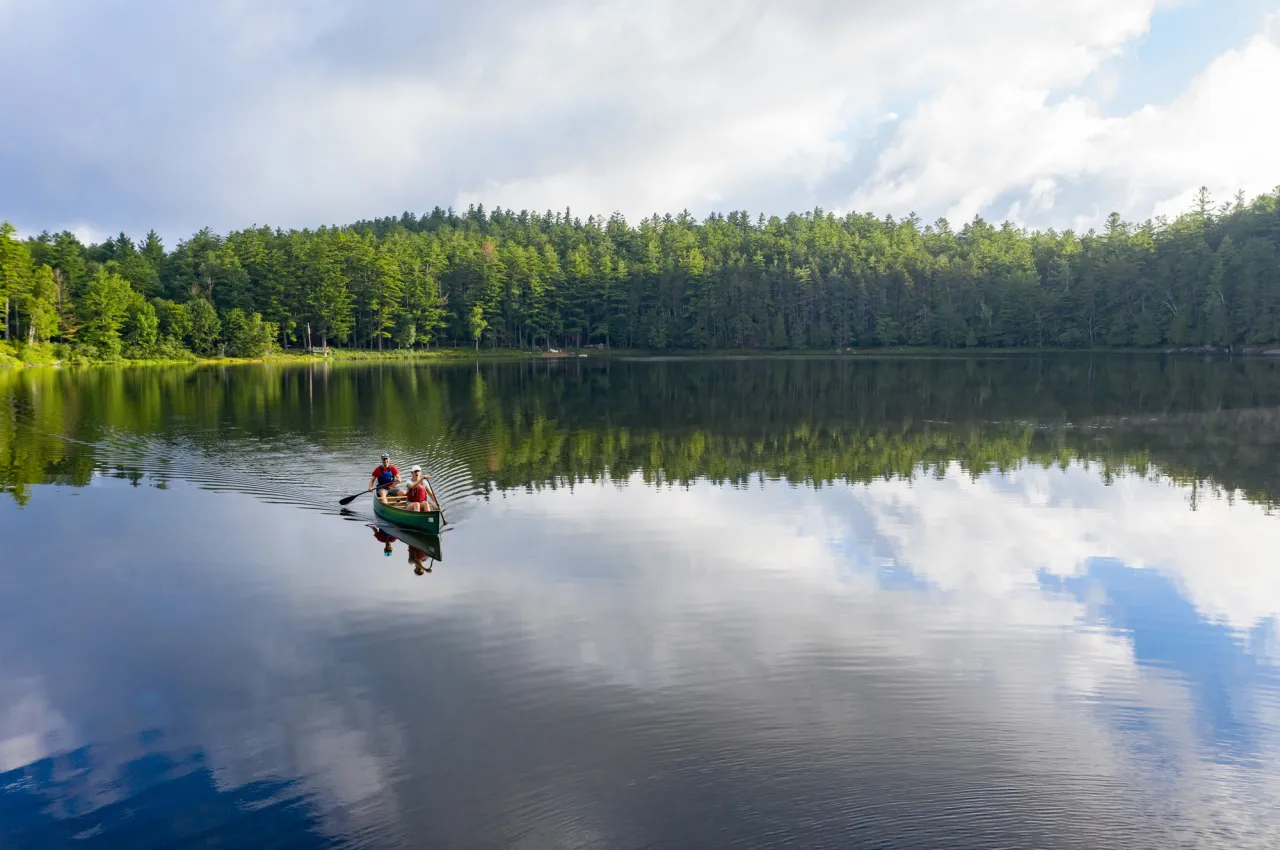 Two people paddle a red canoe across a calm pond surrounded by trees.