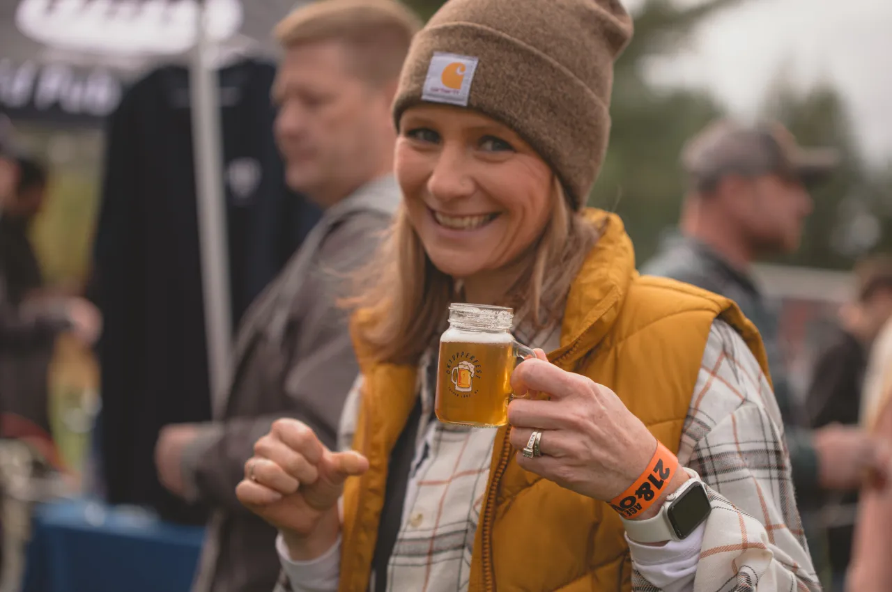 A woman in an orange puffer vest holds up a tiny beer. 