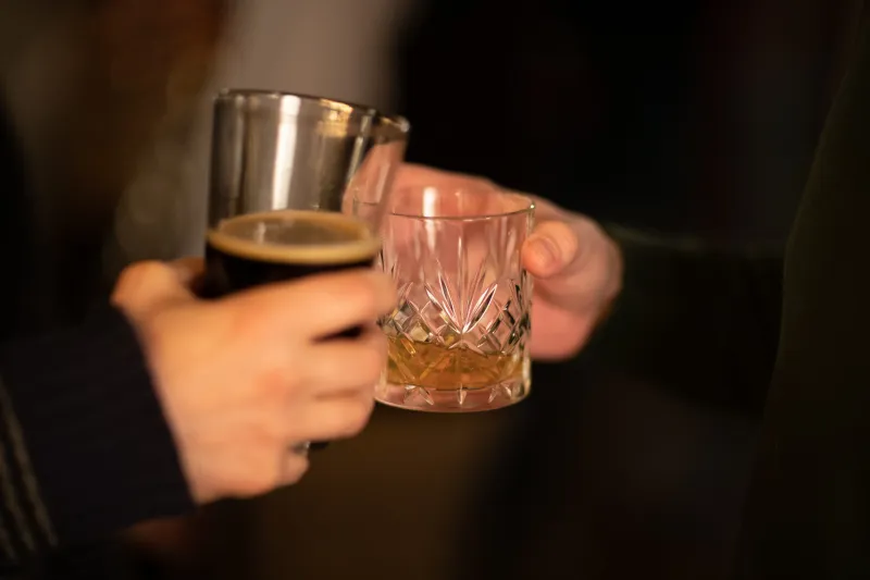 A woman holding a beer and a man holding whiskey glasses clinking together.