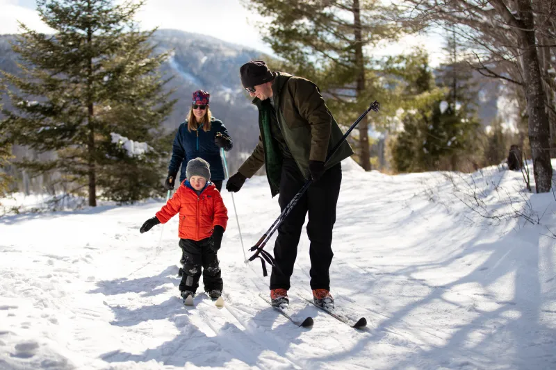 A mother and father teaching their son to cross country ski on a groomed trail.