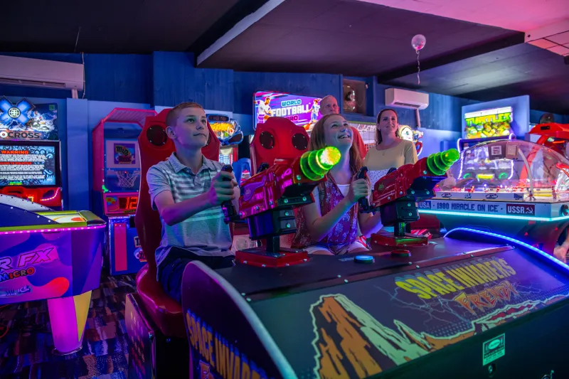 Two smiling children aim plastic laser guns in a brightly colored arcade surrounded by elaborate games.