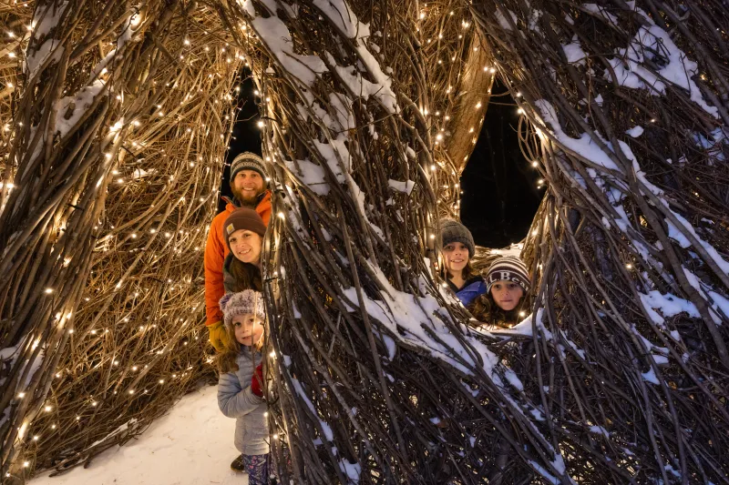 Two adults and three children peek out from behind a large outdoor sculpture made of twigs, decorated with white Christmas lights and a dusting of snow.