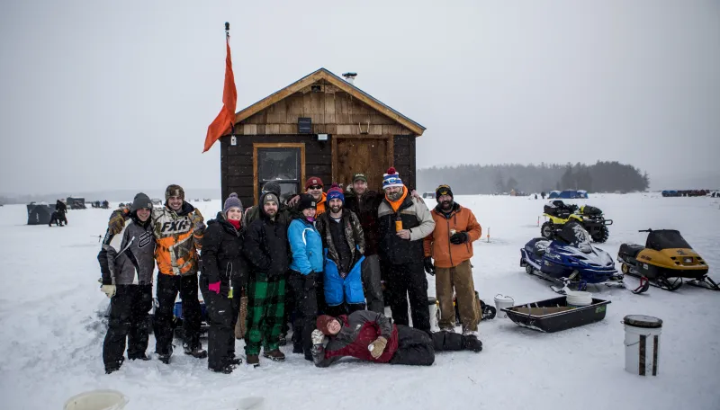 A group of ice fishermen and fisherwomen smile in front of a shack on the ice. Snowmobiles and fishing tents dot the snowy ice.