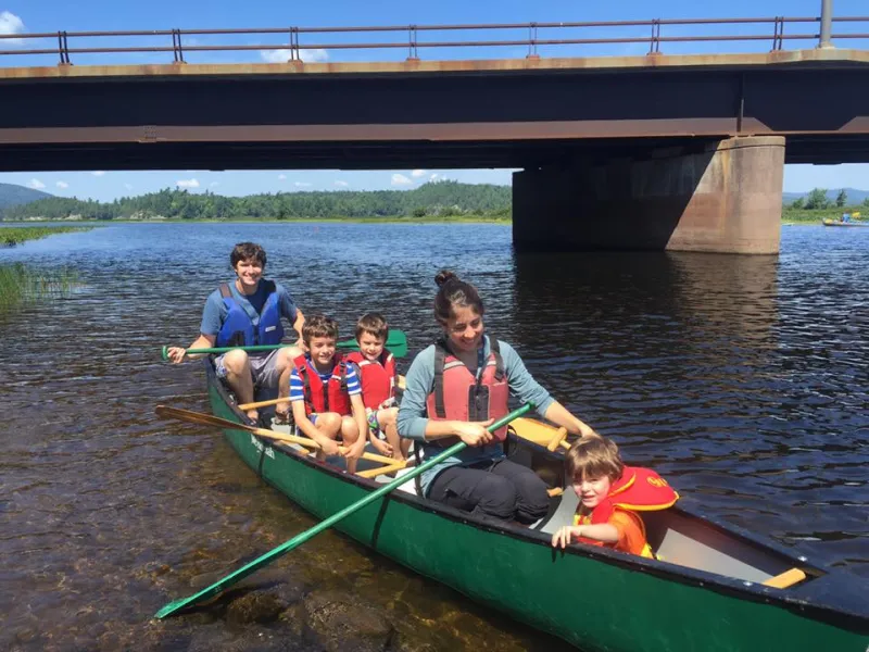 A family of two adults and three young children paddle in a canoe along a lake