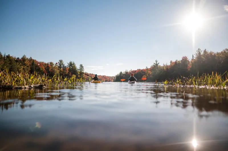 Two women paddle kayakas along a lake with fall foliage along the shoreline