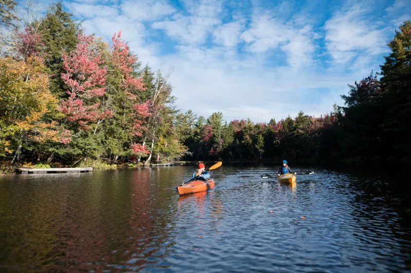 Two women paddle kayakas along a lake with fall foliage along the shoreline