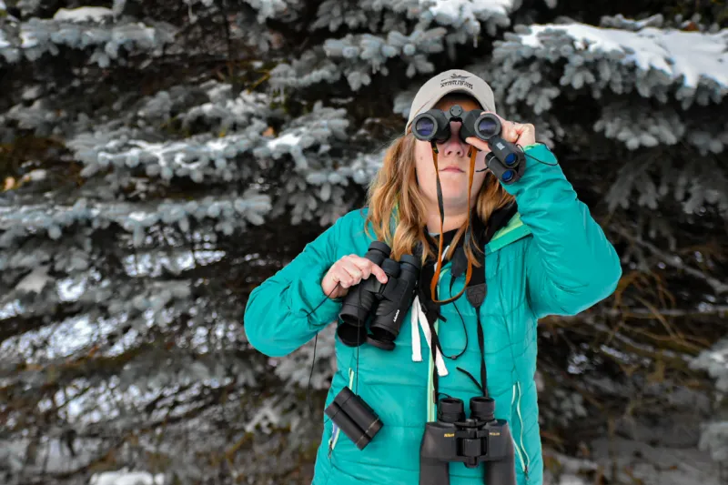 A woman in a green jacket and tan hat looks through a pair of binoculars while holding 4 other pairs. This is the definition of overkill.