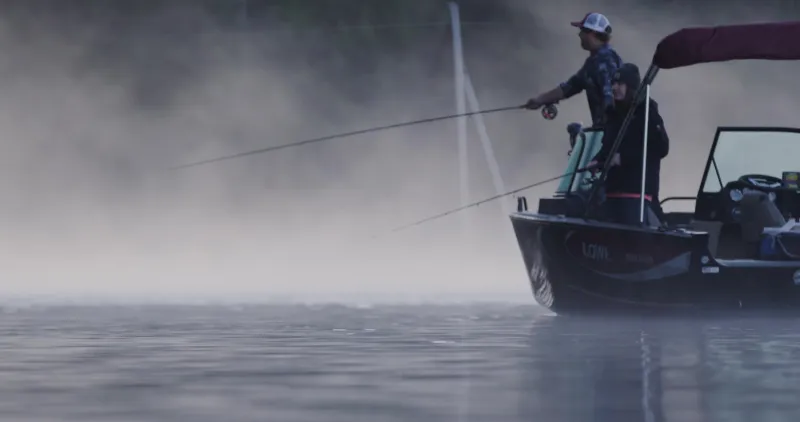 a man and woman fish from a boat on the water with morning fog