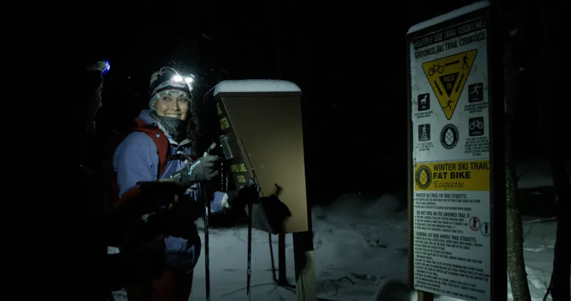 A woman wearing a headlamp smiles at a friend at a skiing trailhead