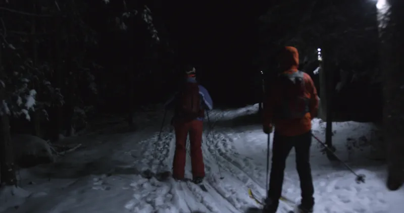 Two people ski downhill past lights attached to trees