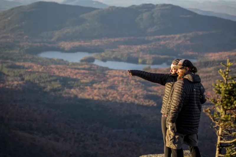 A couple on a mountain summit point off camera, with forest and lakes in the background.