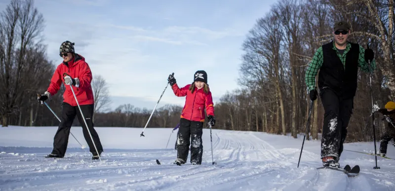 A family of three cross country skies with a young girl in the middle of the trail smiling.